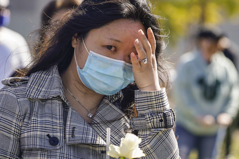 An Asian woman in a face mask with a white flower wiping a tear from her eye