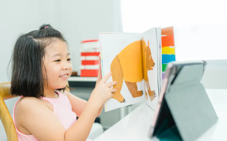 A child holds a picture book in front of a computer.