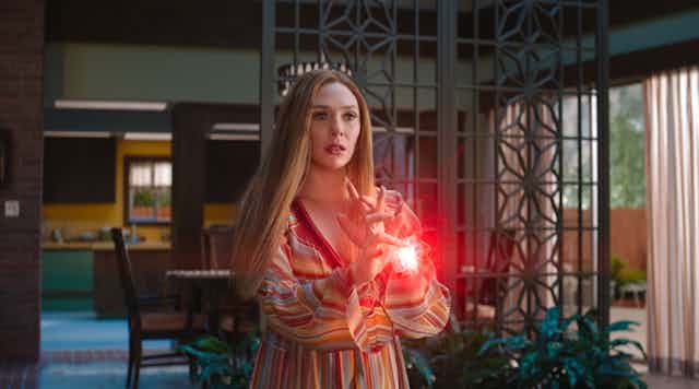 Young white woman with long red hair wearing a striped shirt, conjuring a ball of mystical red light between her hands. Still photo from the TV show Wandavision.