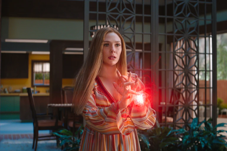 Young white woman with long red hair wearing a striped shirt, conjuring a ball of mystical red light between her hands. Still photo from the TV show Wandavision.