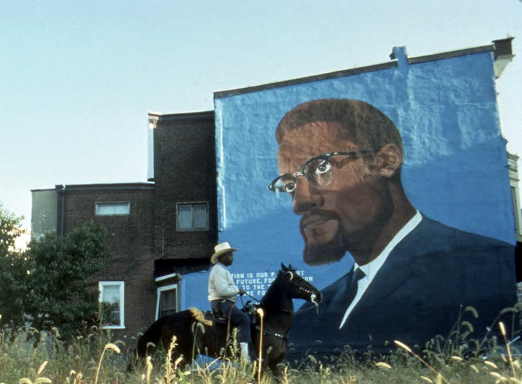 A man rides a horse in front of a mural of Malcolm X.