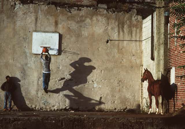 Two boys play basketball as a horse looks on.