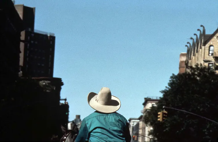 A man wearing a white cowboy hat rides a horse through a city street.