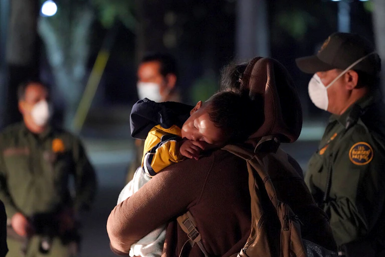 A migrant child sleeps on the shoulder of a woman at an intake area after turning themselves in upon crossing the U.S.-Mexico border, with U.S. border patrol agents standing around them.