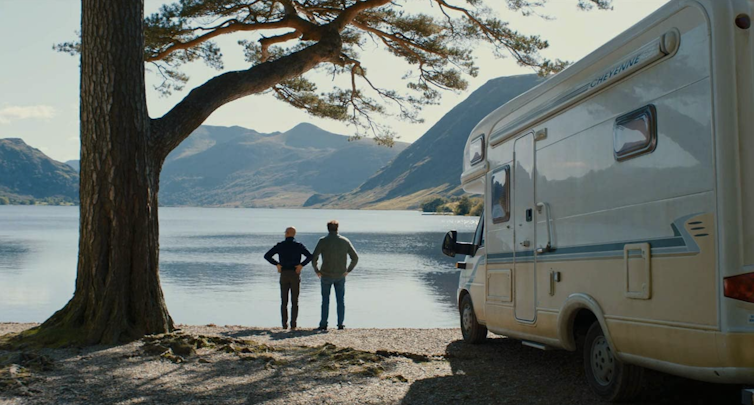 Two men stand by lake with campervan.