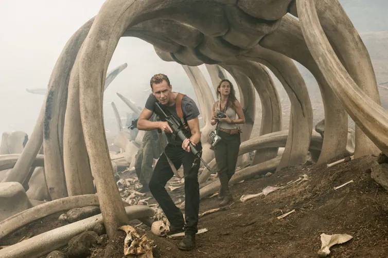 Two people, one with a gun raised and one with a camera search under dinosaur bones.