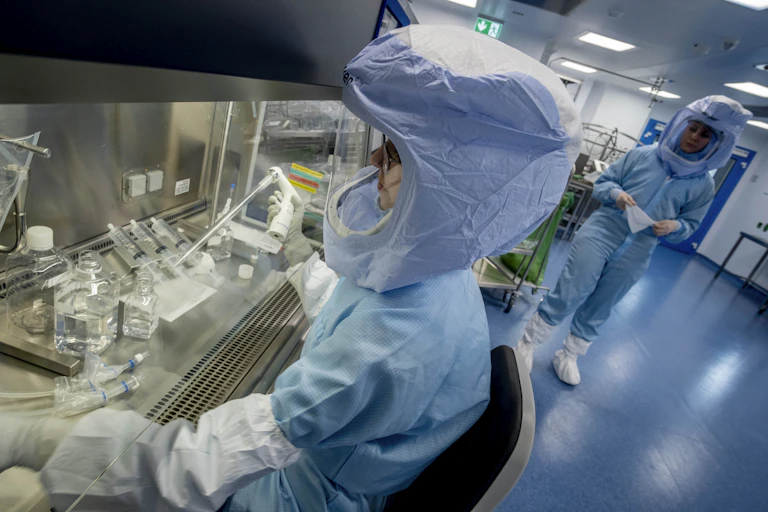 A scientist in a hood and other protective equipment works with glass bottles and equipment.