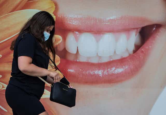 A woman in a face mask walking past a large close-up photo of a smiling mouth
