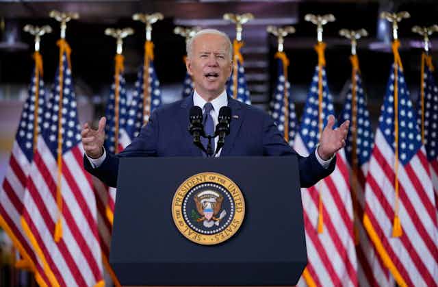 President Joe Biden holds his hands out wide as he gives a speech at a lectern with a row of American flags behind him.