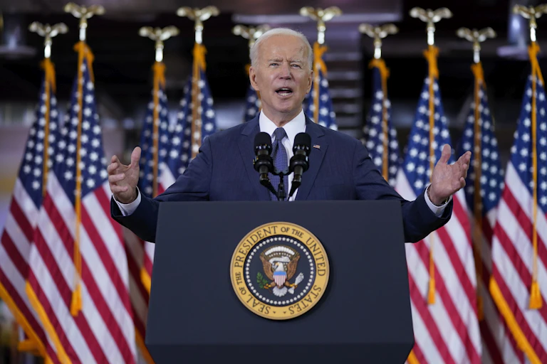 President Joe Biden holds his hands out wide as he gives a speech at a lectern with a row of American flags behind him.