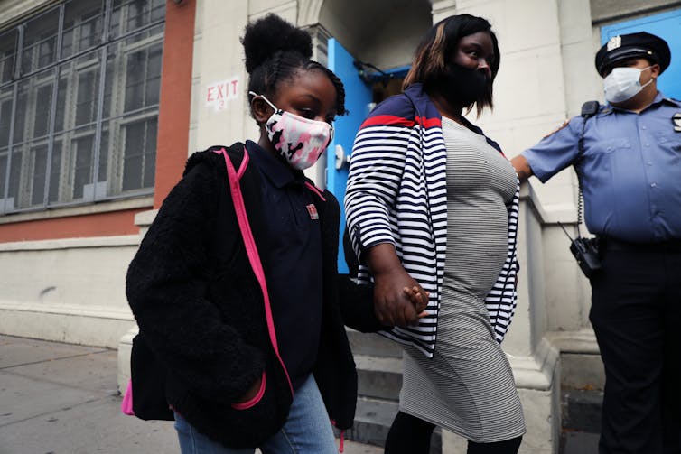 An African American mother and her daughter both wear masks while holding hands walking down the street.