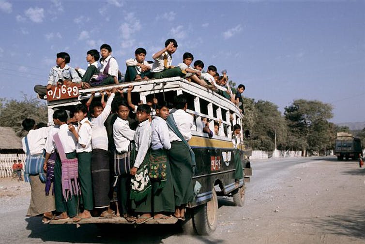 Crianças com blusas brancas e calças verdes estão penduradas do lado de fora de um ônibus muito lotado;  outras crianças sentam em cima do ônibus