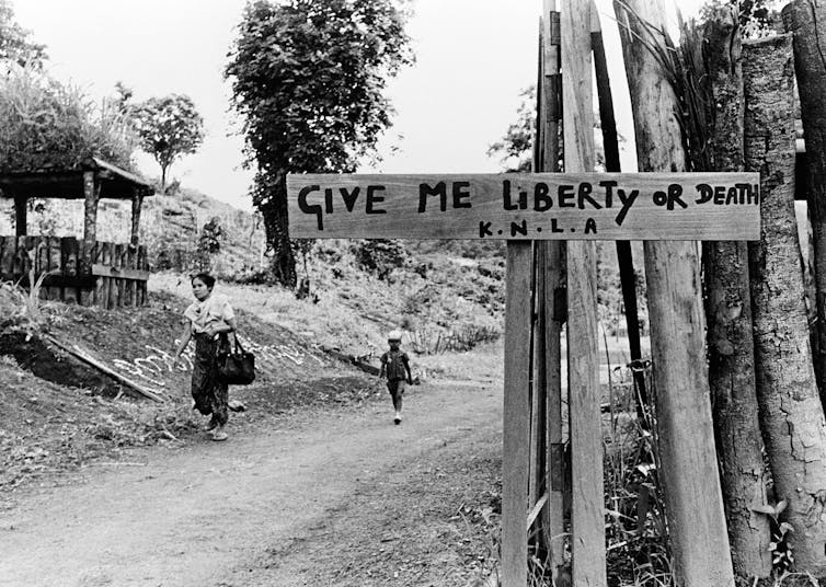 Black-and-white image of a guard hut and a wooden sign reading 'give me liberty or give me death'