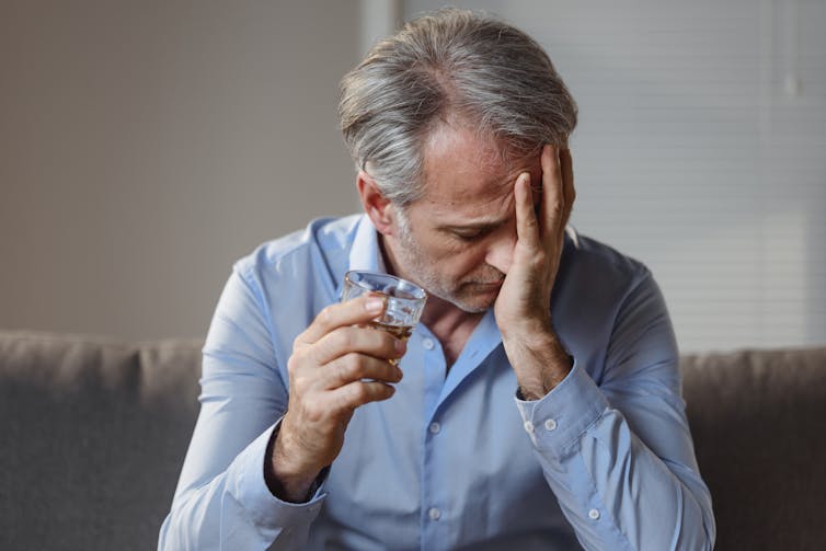 A man sits with a drink glass in one hand and the other hand over his face