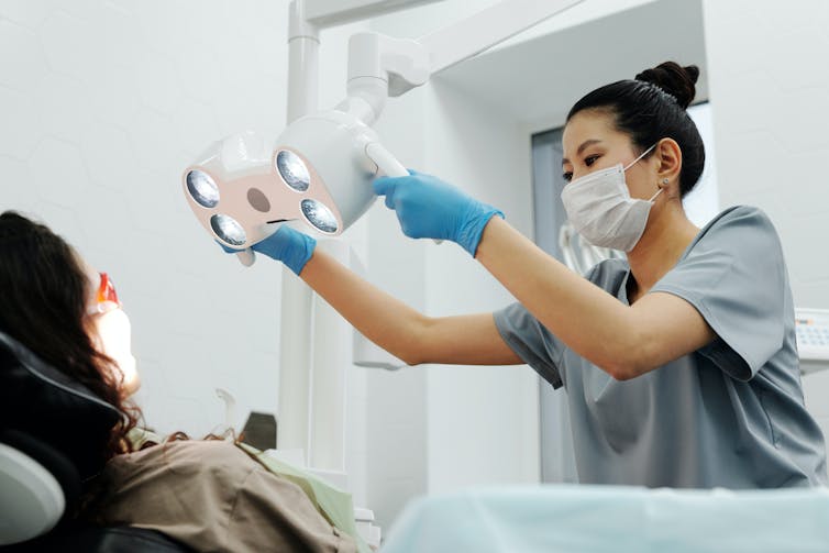 A woman in scrubs and a surgical mask positions a light over a person in a dentist's chair