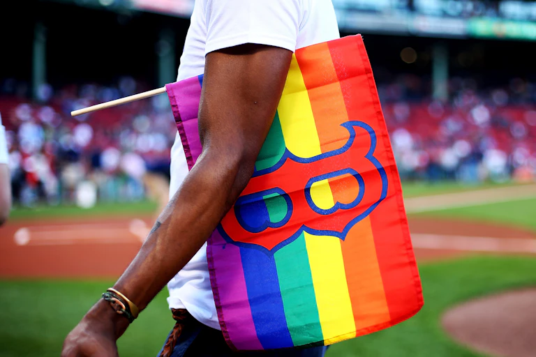 A man with a gay pride flag with a Boston Red Sox logo on it tucked under his arm.