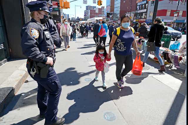 A police officer stands on a sidewalk in a majority Asian neighborhood in New York City as pedestrians walk by.