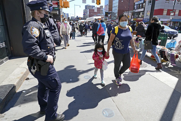 A police officer stands on a sidewalk in a majority Asian neighborhood in New York City as pedestrians walk by.