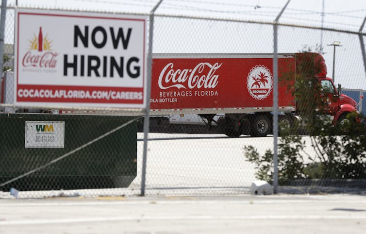 A tractor trailer truck backs into a loading dock at Coca-Cola Beverages Florida past a Now Hiring sign.