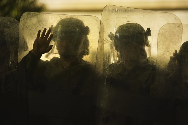Police in riot gear peer through their shields.