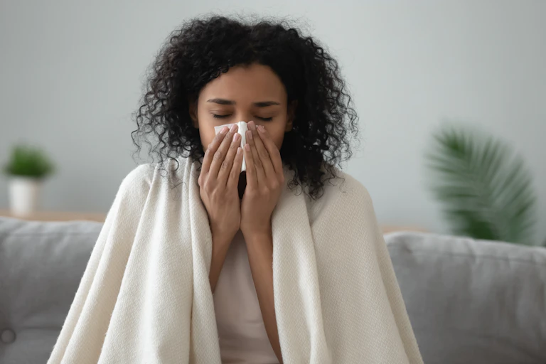 A woman with a blanket over her shoulders blowing her nose.