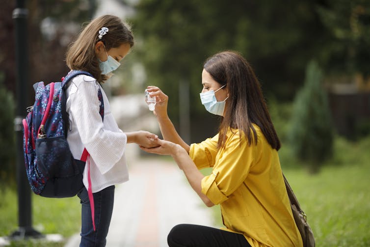masked mother helps masked daughter in backpack with hand sanitizer