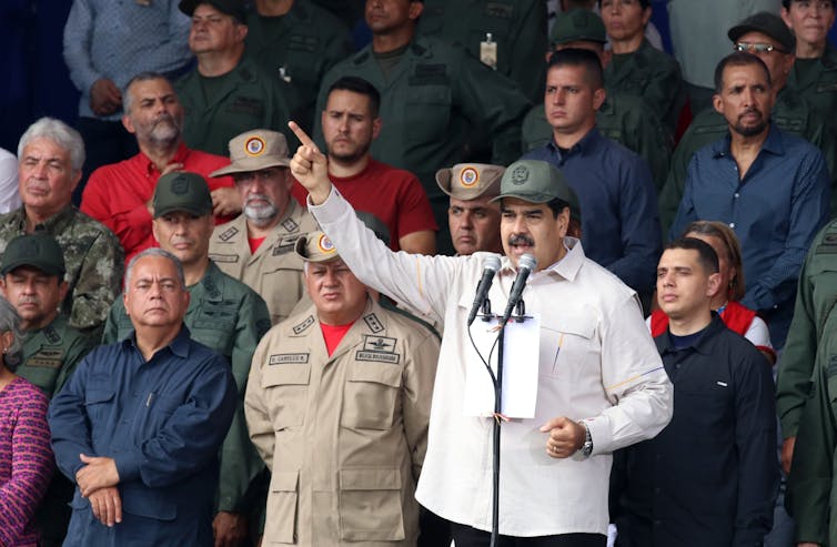 Maduro in a military hat surrounded by soldiers speaks at a microphone with his hand raised