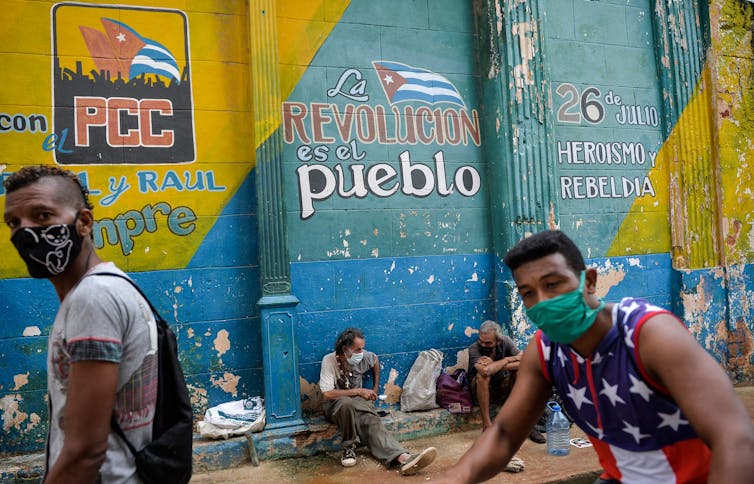 Cubans wearing face masks wait to buy food near a mural about the Cuban Communist Party and the Cuban Revolution.