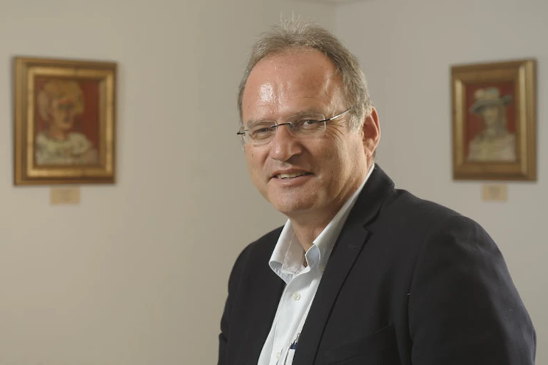A bespectacled, balding man wearing a white shirt and black jacket smiles warmly