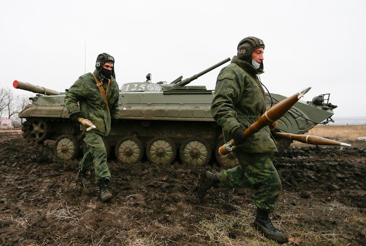 Two men in military uniforms carrying weapons with a Russian-made tank in the background.
