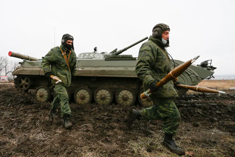 Two men in military uniforms carrying weapons with a Russian-made tank in the background.