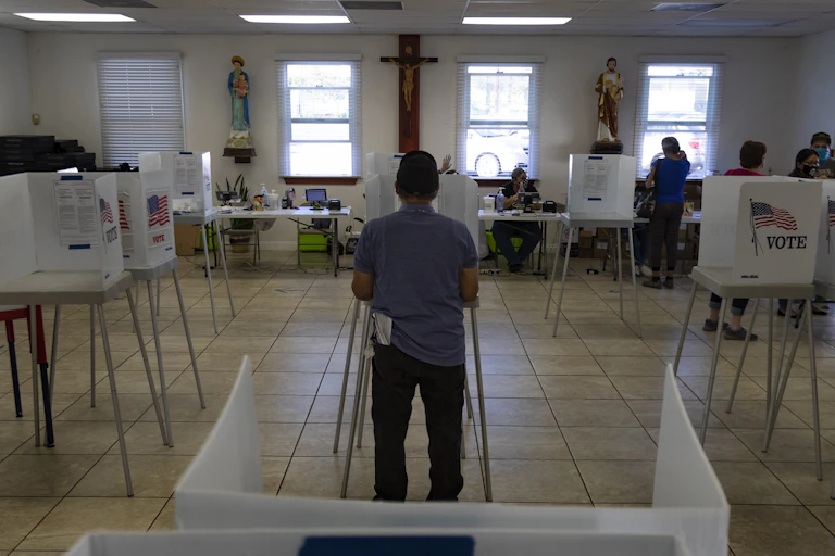 A voter fills out a ballot with Christ on the crucifix hanging on the wall in front of him.