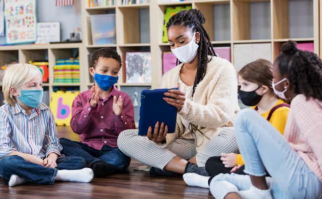 A group of students wearing masks surround a teacher who is also wearing a mask to look at a tablet.
