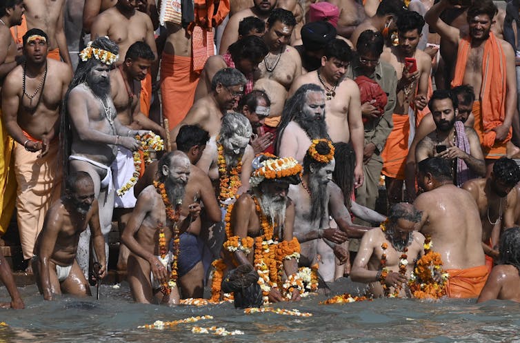 Men are shoulder to shoulder on the edge of the water. Most are naked from the waist up. Many wear flowers and orange clothing.