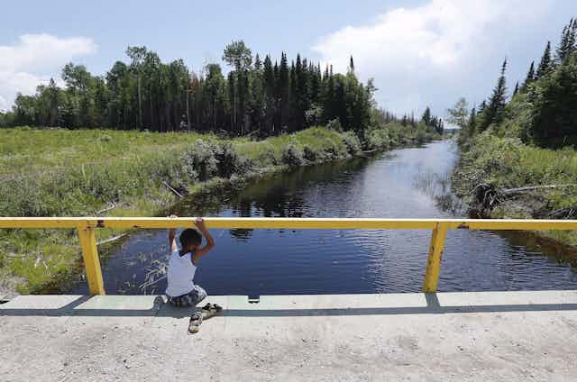 A boy sits on a bridge over a man-made channel in the First Nation of Shoal Lake 40 located in the northern boreal forest, straddling the Manitoba/Ontario border, in June 2015.