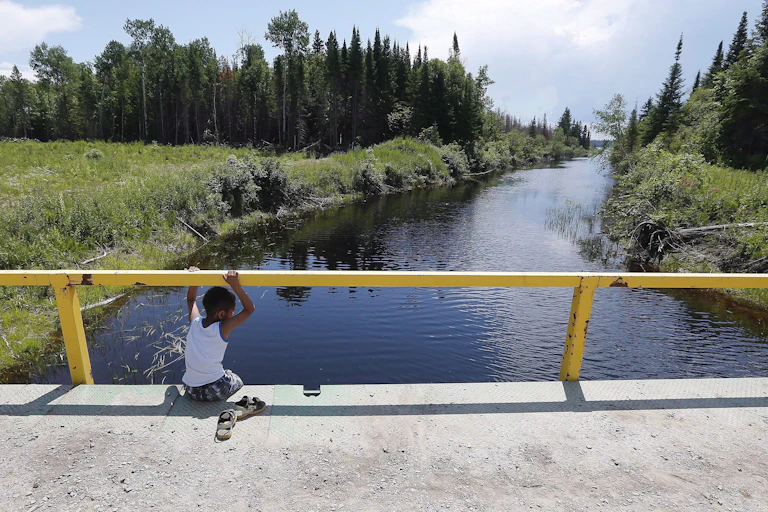 A boy sits on a bridge over a man-made channel in the First Nation of Shoal Lake 40 located in the northern boreal forest, straddling the Manitoba/Ontario border, in June 2015.