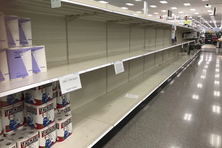 A few packages of paper towels sit on otherwise mostly empty shelves in a store
