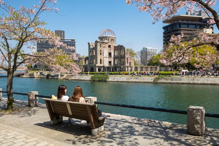 Tourists sitting on bench looking out onto river opposite the Hiroshima Peace Park in Japan
