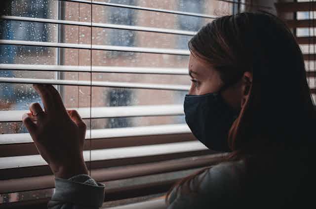 A woman wearing a mask peers out a rainy window.