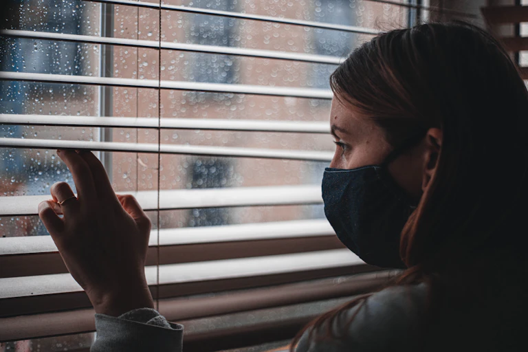 A woman wearing a mask peers out a rainy window.
