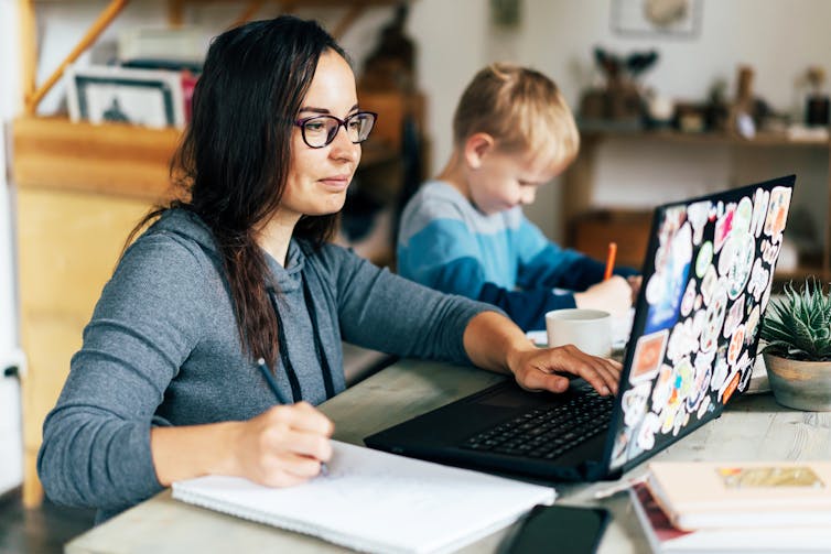 Woman working on laptop while child draws in background.