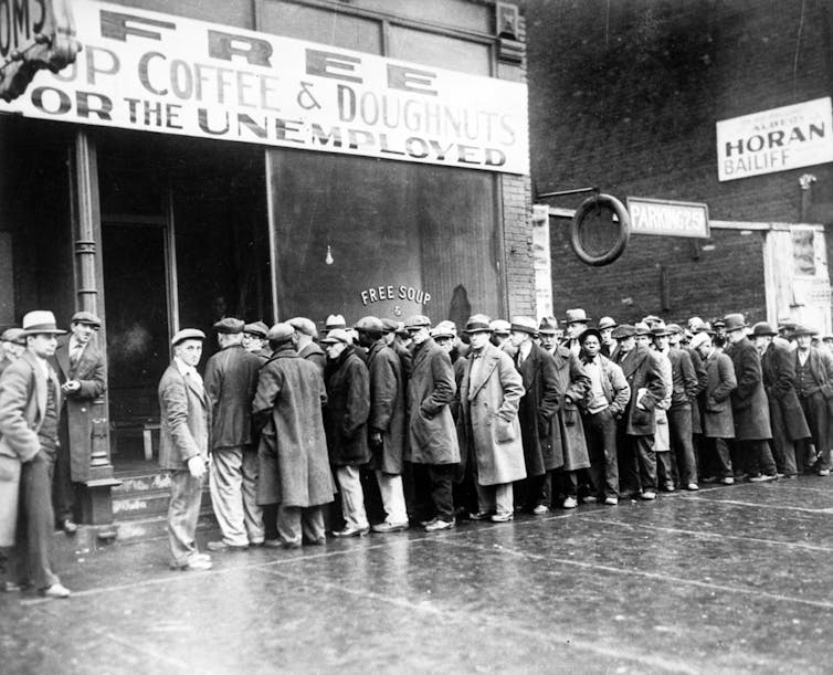 Black and white photo of men in coats and hats queing outside building with sign reading 'Free soup coffee and doughnuts for the unemployed'.