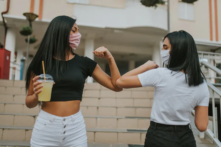 Two women wearing masks greet each other with elbow bumps.