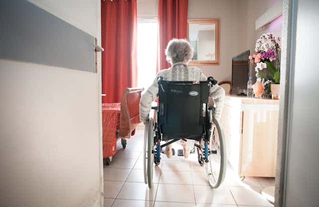 A woman in a wheelchair enters a bedroom at a nursing home.