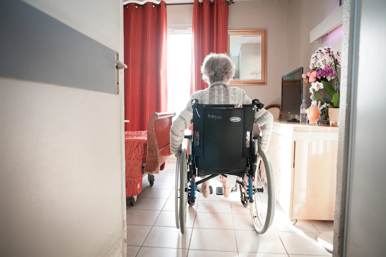 A woman in a wheelchair enters a bedroom at a nursing home.