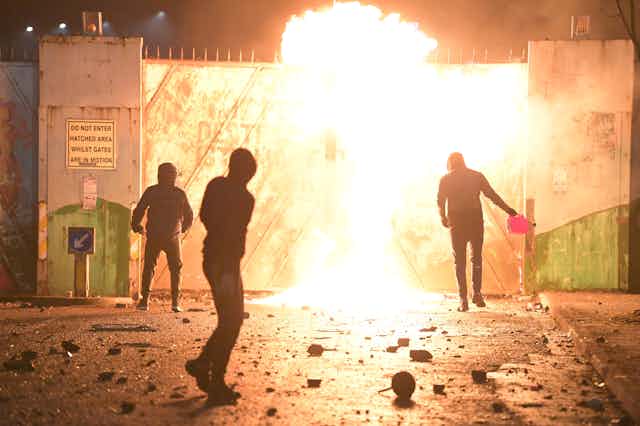 Silhouetted people in hoodies against a burning gate; one person is holding a fuel container