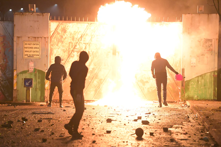 Silhouetted people in hoodies against a burning gate; one person is holding a fuel container
