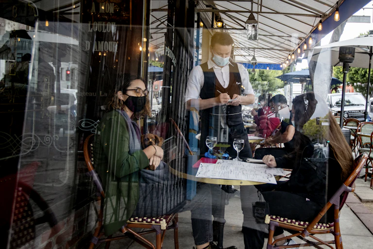 Waiter takes food orders from customers behind a plexiglass at Figaro Bistro in Los Angeles County.