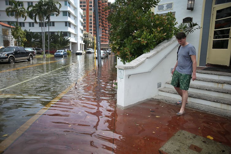 A man wearing flipflops steps onto a **flooded** sidewalk while leaving a hotel.