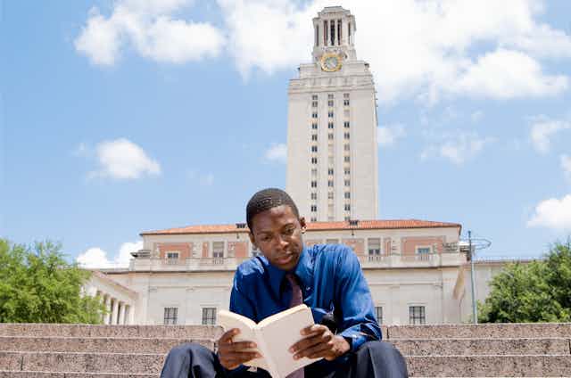 A Black college student reads a book on the steps of a college campus.